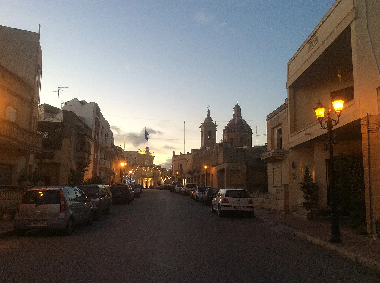 A street in Għaxaq at dusk with the parish church at its end.